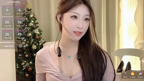 Media: Video of a young Asian woman with long dark hair, wearing a low-cut pink top, seated in front of a decorated Christmas tree. She's using a headset microphone. Background includes a white chair and beige curtains.