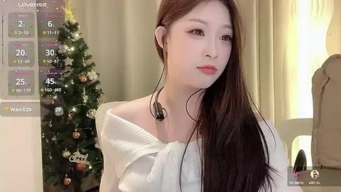 Media: A video of a young East Asian woman with long brown hair, wearing headphones, standing in front of a decorated Christmas tree. She is dressed in a white off-shoulder top.