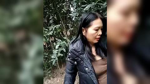 Media: Video of an Asian woman with long black hair and a black puffer jacket, walking in a dense, green forest.