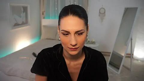 Media: Video of a woman with dark hair, fair skin, and sharp features, wearing a black velvet top, sitting in a softly lit, minimalist room with a large mirror and dreamcatcher decor.