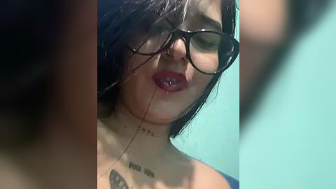 Media: A close-up video of a young woman with dark hair, wearing black-rimmed glasses and red lipstick, leaning against a teal wall. She has a small tattoo on her neck.