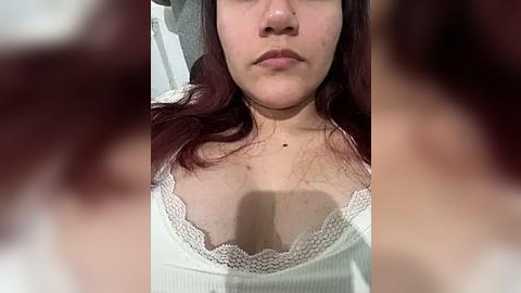 Media: Video of a young woman with fair skin, medium-sized breasts, and long, wavy auburn hair, wearing a white, ribbed tank top with a lace trim. The background is blurred, focusing attention on her face and chest.