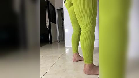 Media: Video of a woman in tight, bright green ribbed leggings, standing barefoot on a tiled floor, viewed from behind. Background shows a white door and a black coat hanging in a hallway.